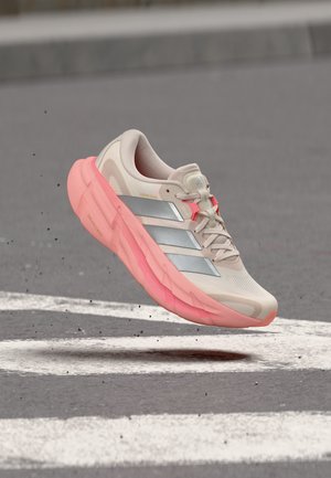 Chaussure de course Adidas beige clair et rose avec des bandes argentées, flottant au-dessus d'une surface asphaltée avec des lignes blanches peintes.