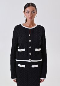 Cardigan nero a maglia con motivo a trecce, con rifiniture bianche, dotato di chiusura frontale a bottoni, due tasche anteriori e grandi bottoni decorativi. Tessuto texture.