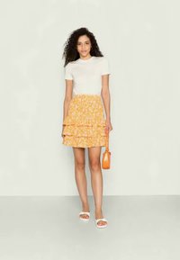 Blusa branca de manga curta combinada com uma saia amarela floral em camadas. Apresenta babados, e a modelo segura uma bolsa laranja. Sandálias brancas completam o look.