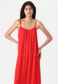 Robe maxi rouge sans manches avec un décolleté froncé et une texture fluide, présentant des fines bretelles et une coupe décontractée.