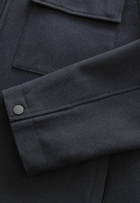 Cappotto blu navy in misto lana con polsini piegati, bottone nero opaco e tasca a toppa, che mette in risalto una morbida texture e un design su misura.