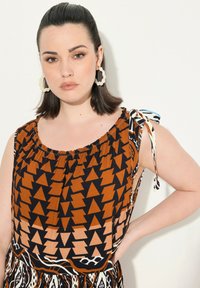 Haut sans manches à motifs en orange et noir avec des designs géométriques ; présente un col froncé, des accents de nœud sur les bretelles et une texture douce.