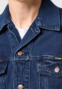 Gros plan d'une personne portant une veste Wrangler en denim bleu avec des boutons en cuivre, par-dessus une chemise blanche, montrant le col et la partie supérieure de la poitrine.