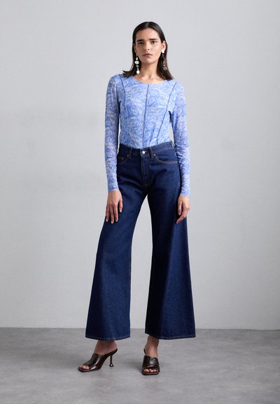 Φωτεινό μπλε πουκάμισο με μακριά μανίκια και λουλούδινη σχέδιο, συνδυασμένο με ψηλόμεσες σκούρες τζιν παντελόνες με φαρδιά γραμμή και καφέ ψηλές παντόφλες.