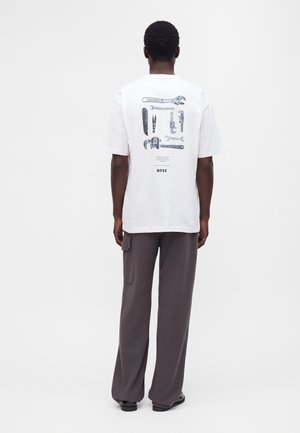 Homme en T-shirt blanc avec des illustrations d'outils noires et grises et un pantalon gris, debout, dos tourné, sur fond blanc.