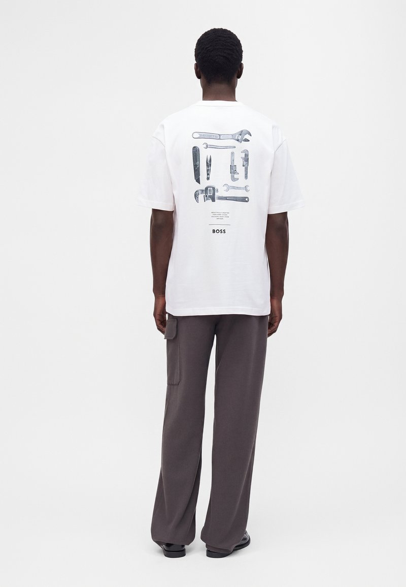 Homme en T-shirt blanc avec des illustrations d'outils noires et grises et un pantalon gris, debout, dos tourné, sur fond blanc.