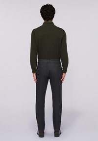 Uomo in piedi di spalle, indossa una camicia a maniche lunghe verde scuro, pantaloni slim-fit grigio scuro e scarpe nere su uno sfondo semplice.