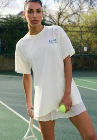 Vit oversized t-shirt med blå "CLUB"-logotyp och veckad nederkant. Modellen håller i ett tennisracket och boll, stående på en grön tennisbana.
