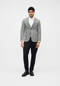 Νέος άνδρας που στέκεται, φορώντας γκρι καρό σακάκι, λευκό πουκάμισο, μαύρο παντελόνι και μαύρα επίσημα παπούτσια, σε απλό λευκό φόντο.