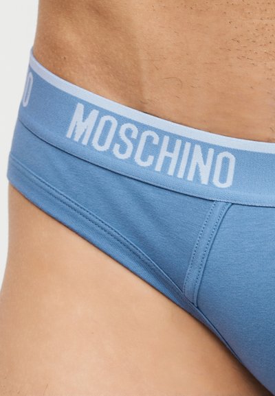 Κοντινό πλάνο ενός ατόμου που φοράει μπλε εσώρουχα με ανοιχτό μπλε ελαστική ζώνη με την επιγραφή "MOSCHINO".