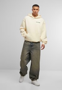Sweat à capuche crème avec poche avant et texte "anomalie" en vert, associé à un jean délavé à coupe large et des baskets blanches.