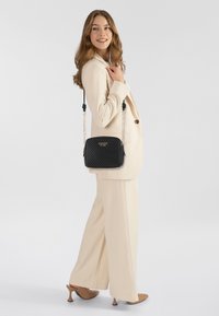 Borsa a tracolla nera trapuntata con branding dorato, abbinata a un completo crema. Presenta dettagli con catena e cinturino regolabile, texture liscia.