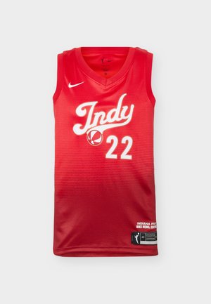 Fiatal férfi göndör hajjal, piros Indiana Fever kosárlabdamezzel, amelyen a 22-es szám látható, és fekete Nike rövidnadrágban, fehér háttér előtt áll.