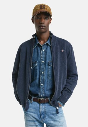 Giovane uomo con un cappello marrone, camicia di jeans blu, giacca blu navy con zip, jeans azzurri chiari e una cintura marrone scuro, con le mani in tasca.