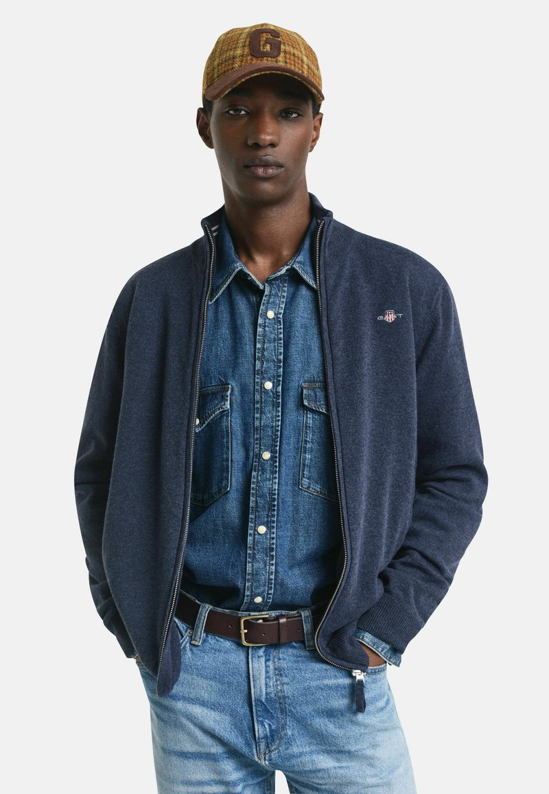 Ung man som bär en brun keps, en blå denimskjorta, en marinblå zip-up-jacka, ljusblå jeans och ett mörkbrunt bälte med händerna i fickorna.