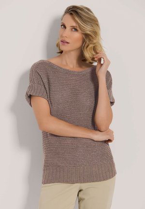 Frau mit blonden Haaren, die ein mauvefarbenes kurzärmliges Strickoberteil und beige Hose trägt, posiert mit einer Hand, die ihr Haar berührt, und der anderen Hand, die verschränkt ist.