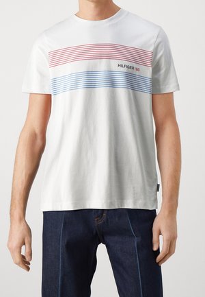 Mężczyzna w białej koszulce z krótkim rękawem z czerwonymi i niebieskimi poziomymi paskami oraz napisem "HILFIGER '85", w zestawieniu z ciemnoniebieskimi dżinsami.