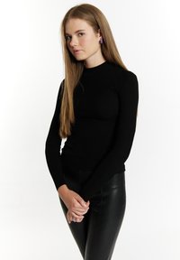 Top a maniche lunghe nero a coste con colletto alto, abbinato a pantaloni in pelle neri eleganti. La modella ha i capelli lunghi e lisci e delle orecchini viola.