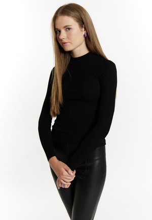 Schwarzes, geripptes Langarmoberteil mit hohem Kragen, kombiniert mit eleganten schwarzen Lederhosen. Das Model hat glattes, langes Haar und trägt lila Ohrringe.