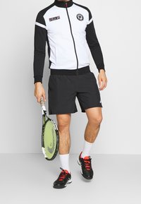 Giacca da tuta nera e bianca con zip, pantaloni corti neri, calze bianche e scarpe da tennis nere, mentre tiene in mano una racchetta da tennis verde e nera.