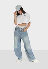 Polo bianco cropped con dettaglio a forma di cuore blu, abbinato a jeans denim leggermente larghi di un blu chiaro e a un cappellino blu chiaro. Le sneakers completano il look.