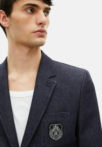 Marineblauer Denim-Blazer mit einschlägigen Revers, strukturiertem Stoff und einer Brusttasche mit einem silbernen Emblem. Weißes Unterhemd darunter sichtbar.