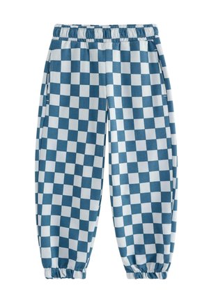 Ung dreng iført en cremefarvet sweatshirt med brune og blå ærmer, blå- og hvide ternede bukser og blå sneakers, stående og smilende.