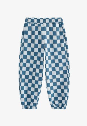 Ung dreng iført en cremefarvet sweatshirt med brune og blå ærmer, blå- og hvide ternede bukser og blå sneakers, stående og smilende.