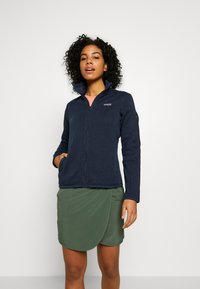Giacca in fleece blu navy con colletto alto, zip frontale e tasche laterali, abbinata a una gonna avvolgente verde. Texture liscia e design aderente.