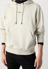 Béžová mikina s kapucí, s klokaní kapsou, žebrovanými manžetami a šňůrkami s černými akcenty. Logo "BOSS" potištěné na hrudi.