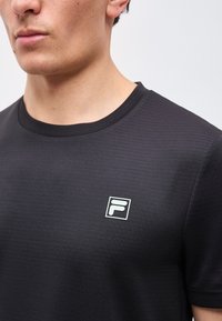 Νεαρός άνδρας φοράει μαύρο t-shirt με στρογγυλή λαιμόκοψη και μικρό λευκό λογότυπο "F" στην αριστερή πλευρά του στήθους, μπροστά σε απλό φόντο.