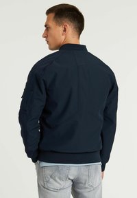 Giacca bomber navy con finitura liscia, colletto e polsini a coste, e tasca con zip sulla manica. Presenta un design aderente e cuciture piatte.