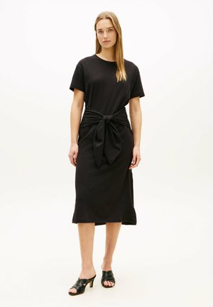 Kvinde iført en sort midi-kjole med korte ærmer og bindebånd i taljen, kombineret med sorte hæl-sandaler, stående foran en ensfarvet baggrund.