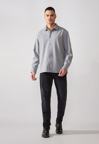 Camicia grigia con bottoni, maniche lunghe e colletto, realizzata in tessuto liscio. Abbinata a pantaloni neri e scarpe nere. Design semplice.