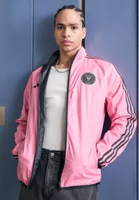Giacca a vento rosa con dettagli neri e logo, dotata di chiusura lampo frontale, tasche laterali e tre strisce nere sulle maniche.