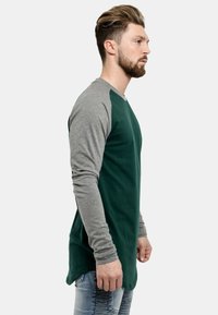 Majica dugih rukava s zelenim tijelom i sivim raglan rukavima, izrađena od mekanog pamučnog materijala, s zakrivljenim rubom i minimalnim detaljima.