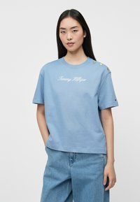 Gaiši zils kokvilnas T-krekls ar īsām piedurknēm, apaļu kaklu un izšūtu "Tommy Hilfiger" tekstu; uz viena pleca ir zelta pogu akcenti.