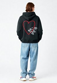 Felpa nera con ricamo a forma di cuore rosso e spille di sicurezza bianche sul retro. Abbinata a jeans boyfriend blu larghi e scarpe sportive multi-colore.