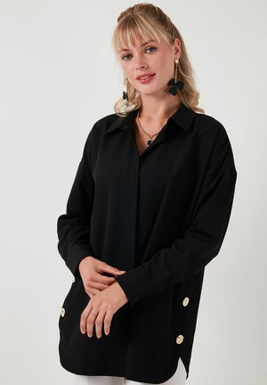 Fekete, hosszú ujjú ing, galléros nyakkal, amely oldalain nagy aranyszínű gombok találhatók, és laza szabással rendelkezik. Simás anyagú textúra.