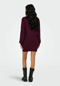 Donna con lunghi capelli neri, che indossa un vestito di maglia viola e stivali neri con fibbia, in piedi di spalle contro uno sfondo bianco semplice.