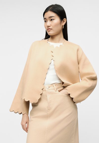 Vrouw draagt een beige jas met een gekartelde rand over een wit shirt en een beige hoge taille rok, met één hand in de zak, kijkt naar de zijkant.