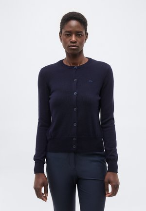 Frau trägt eine marineblaue, geknöpfte Strickjacke und passende Hose, steht vor einem schlichten weißen Hintergrund und blickt nach vorne.