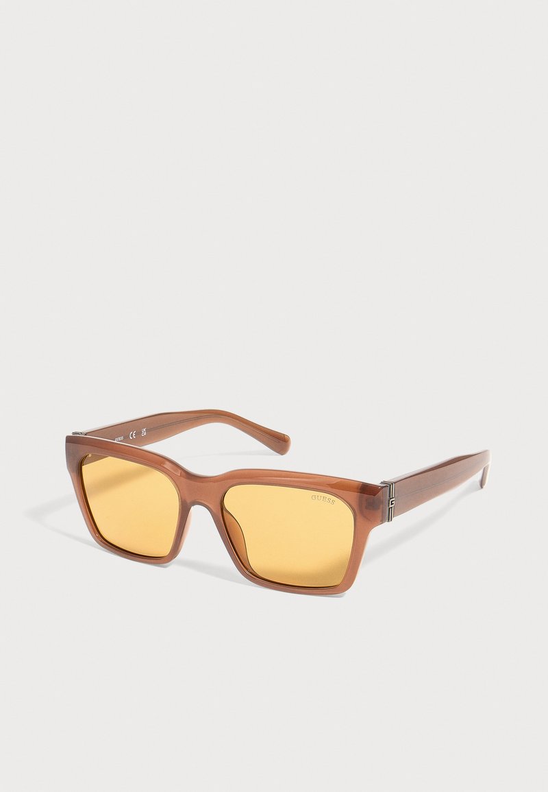 Lunettes de soleil rectangulaires avec monture translucide marron, verres jaunes et détails métalliques sur les branches, posées sur un fond blanc.