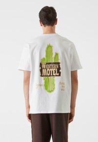Bijela pamučna majica s grafikom zelenog kaktusa i natpisom "WESTERN MOTEL" u vintage stilu na leđima, s dodatnim frazama ispod.