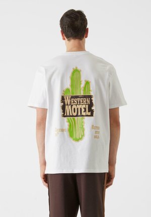 Balta medvilninė marškinėliai su žaliu kaktusu ir užrašu "WESTERN MOTEL" vintage stiliumi ant nugaros, su papildomais posakiais apačioje.