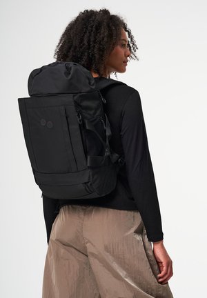 Person mit lockigem Haar, der einen großen schwarzen Rucksack, ein schwarzes Langarmshirt und beige Hosen trägt, seitlich vor einem weißen Hintergrund steht.