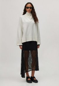 Witte langomliggende gebreide trui met een colkraag, gecombineerd met een zwarte kanten rok met een doorzichtige ontwerp en een asymmetrische zoom. Zwarte schoenen.