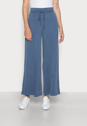 Zijdebroek met wijde pijpen in een zachte, lichte blauwe stof. Voorzien van een trekkoord in de taille en reikt tot de enkel, gecombineerd met witte sneakers.