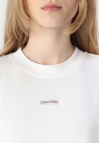 Hvit bomulls t-skjorte med rund hals og et lite rødt "Calvin Klein" logo-lapp på brystet. Glatt tekstur og rent design.