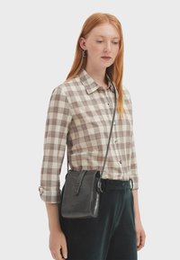 Giovane donna con capelli rossi che indossa una camicia beige a scacchi e una borsa a tracolla in pelle nera, in piedi davanti a uno sfondo semplice.